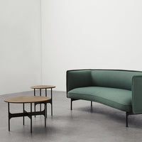 Green curved sofa with black legs and two round, natural wood-topped coffee tables on a light gray floor.