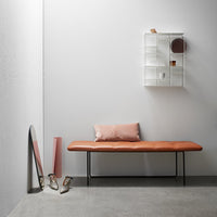 Tan leather bench with pink accent cushion against a minimalist gray wall and geometric white shelving.