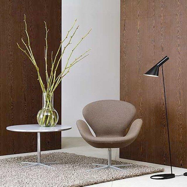 Brown ergonomic chair, white round table, glass vase with green branches, and black floor lamp against a textured brown wall.