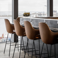 "Modern bar stools in warm brown leather with smooth curves and sleek black metal legs against a scenic backdrop."