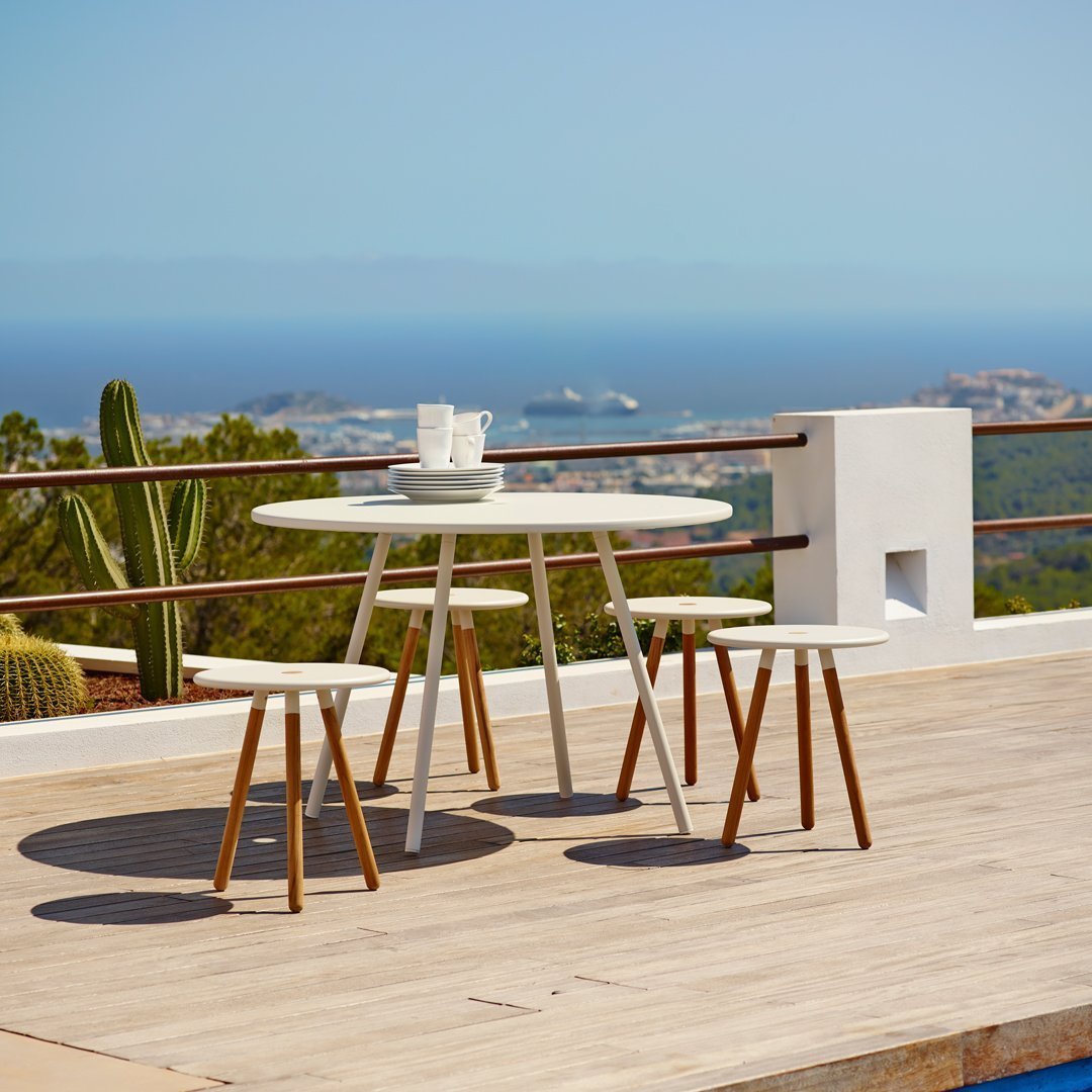Stylish white outdoor table with natural wood legs, set against a serene blue ocean view and lush green plants.
