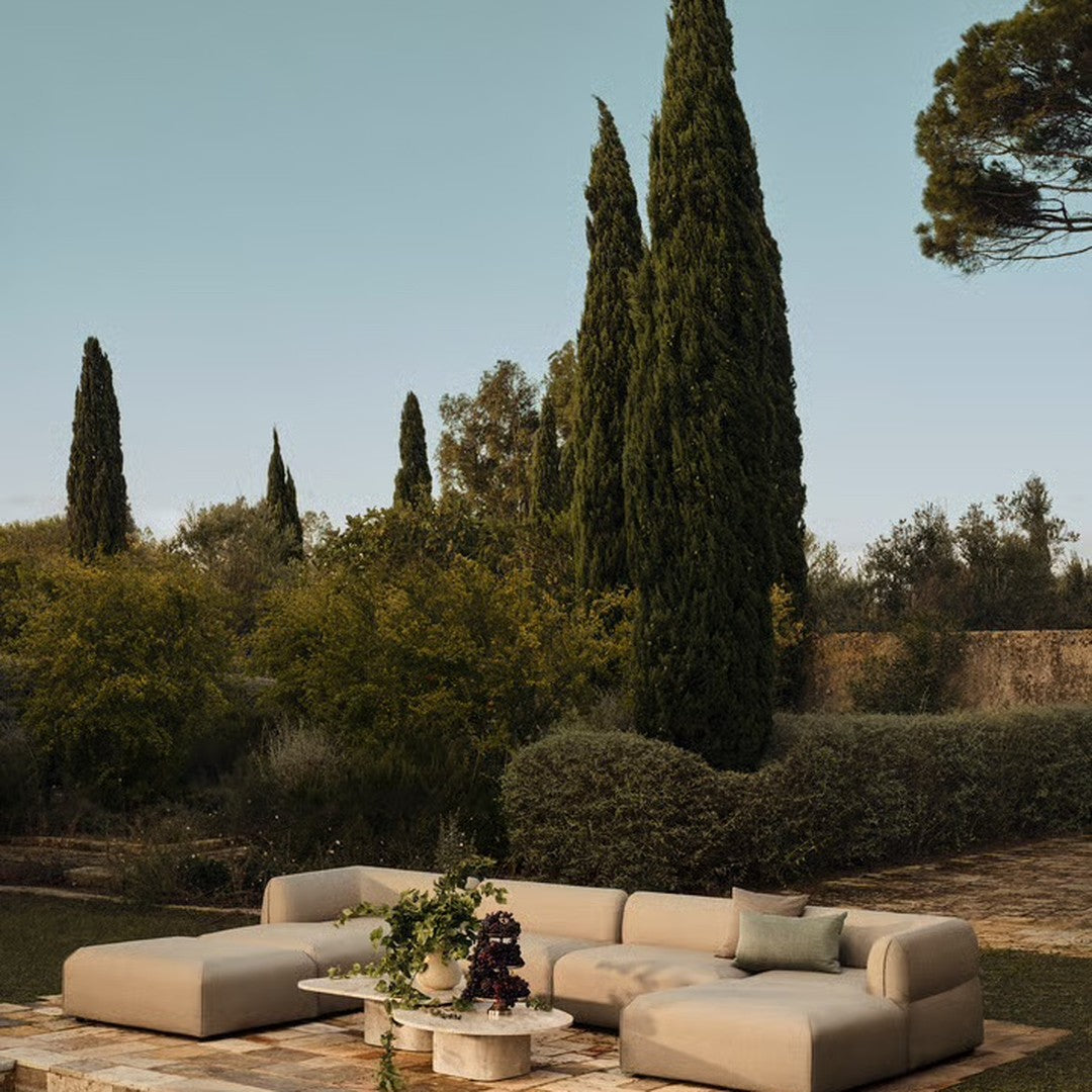 Beige sectional sofa with subtle texture, set against a backdrop of lush greenery and towering trees.