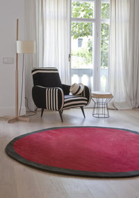 Red round rug with a black border, complementing a black and white striped chair in a bright, airy room.