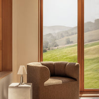 Brown textured upholstery armchair with curved design next to a modern lamp and large windows overlooking a green landscape.