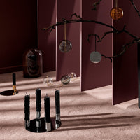 Black candle holder with textured candles, surrounded by warm earthy tones and geometric glass ornaments against a burgundy backdrop.