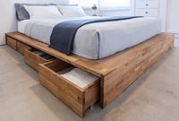 "Light grey bedding with a navy accent blanket on a natural wood platform bed with storage drawers."