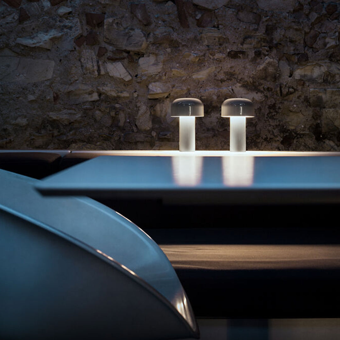 Modern table lamps with a sleek, clear base and soft glow, set against a textured stone wall and dark reflective surfaces.