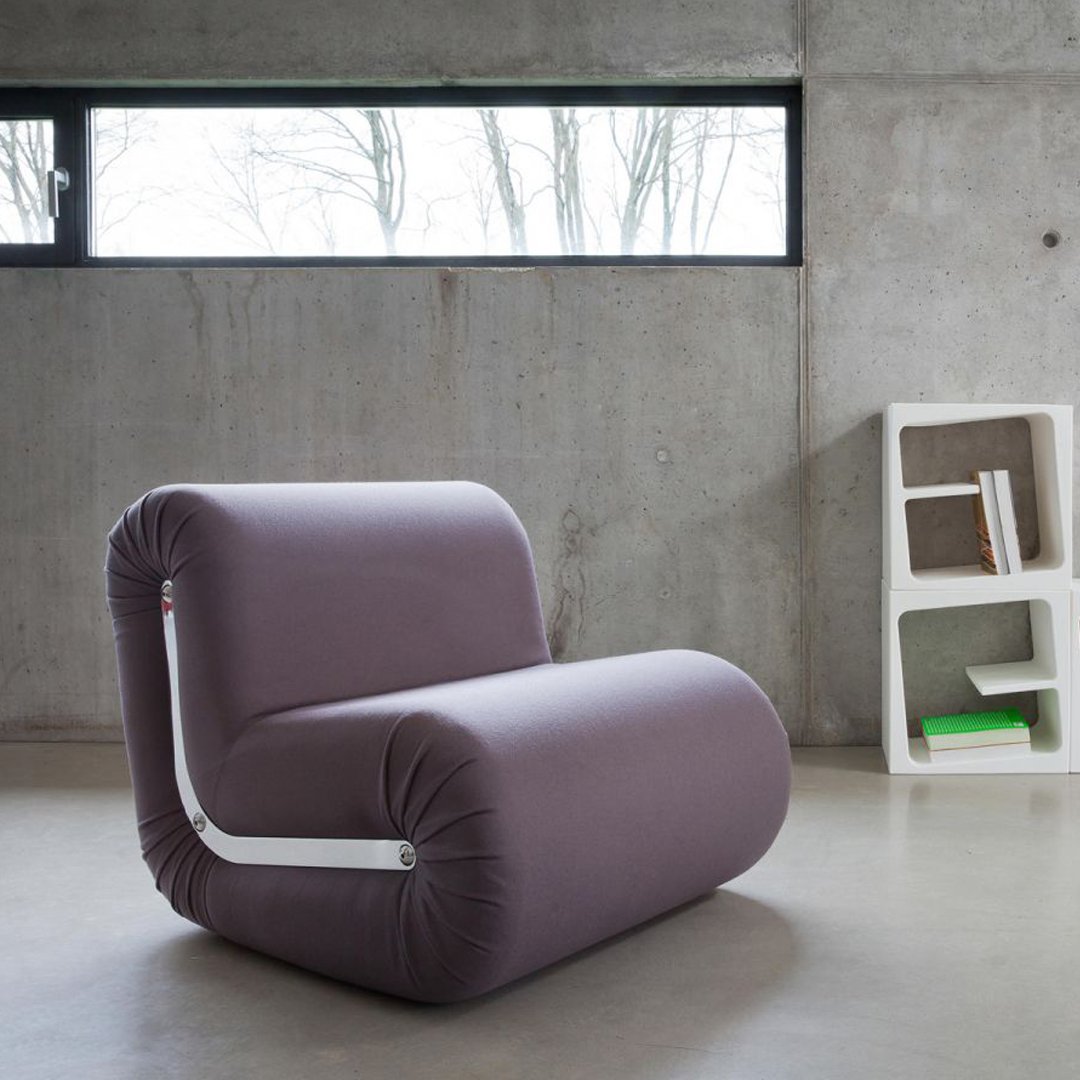 Modern chair in soft purple fabric with smooth, minimalist design and white accent piping against a gray concrete backdrop.