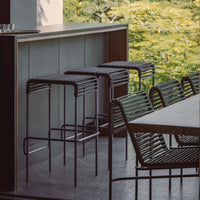 Black woven bar stools alongside a sleek table in an outdoor setting with vibrant green foliage in the background.