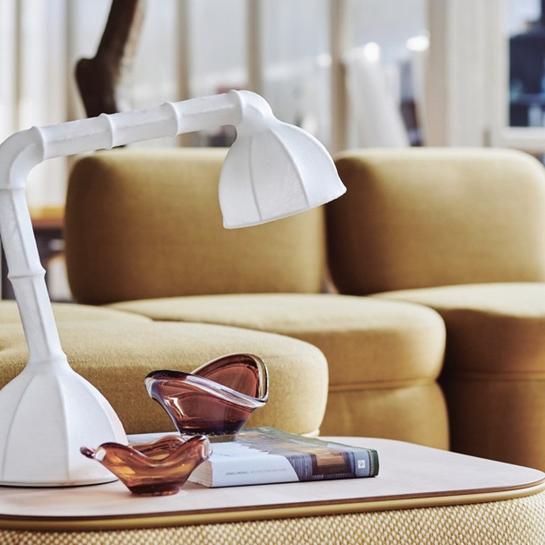 White desk lamp with curved shade, amber glass bowls, and a soft yellow sofa in a modern living room setting.