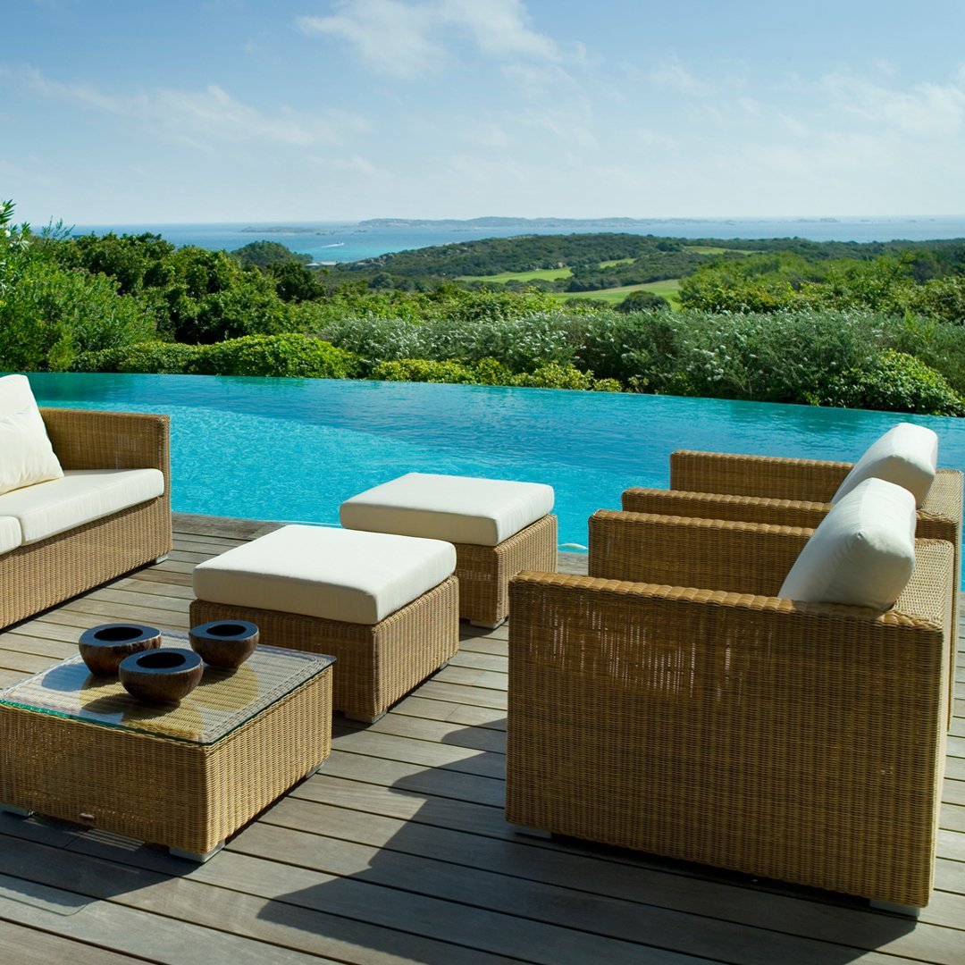 "Light brown wicker patio furniture with cream cushions, set by a turquoise pool and lush green landscape."