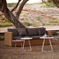 Brown woven outdoor sofa with white and purple accent tables in a natural setting.
