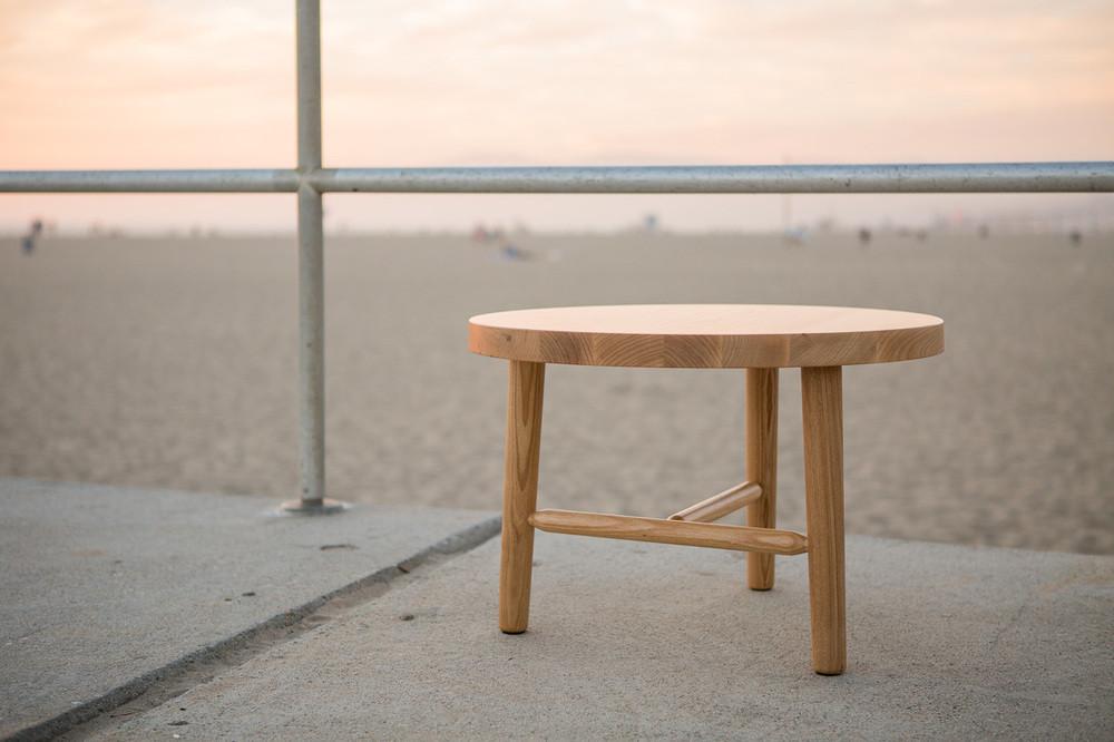 Natural wood round table with a light finish and simple legs, set against a soft pastel sunset background.