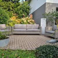 Gray wicker sofa set with neutral cushions, set against a lush green garden backdrop and paved stone patio.