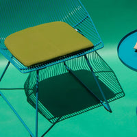 Blue wire chair with a green cushion, casting intricate shadows on a vibrant green background, showcasing modern design.