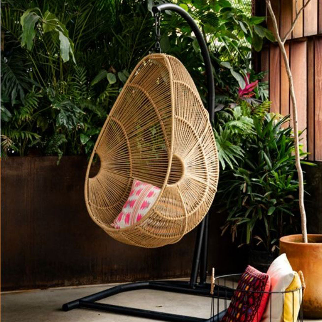 Hanging rattan chair with a natural finish and colorful pink and red patterned cushions, surrounded by lush greenery.