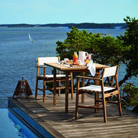 "Outdoor wooden dining set with white cushions, set against a serene blue water backdrop and green trees."