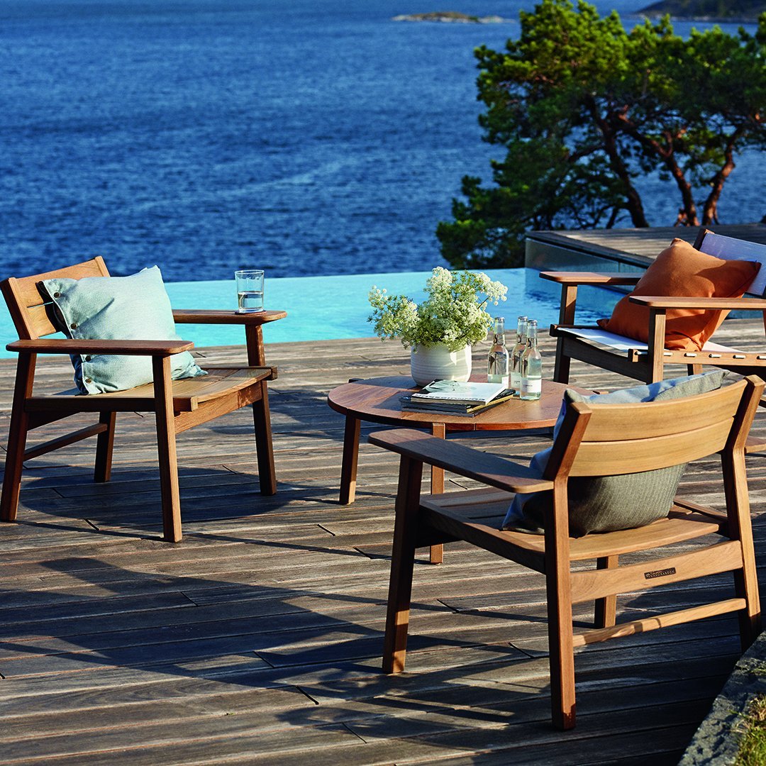 Wooden patio chairs with blue and green cushions, orange accents, set against a tranquil waterfront backdrop.