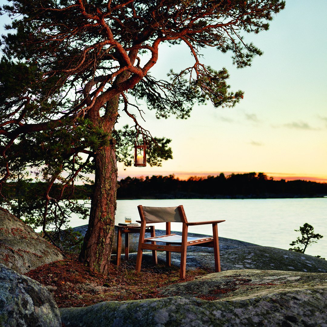 "Wooden chair with beige canvas seat, set against a serene sunset backdrop and lush green tree, creating a natural oasis."