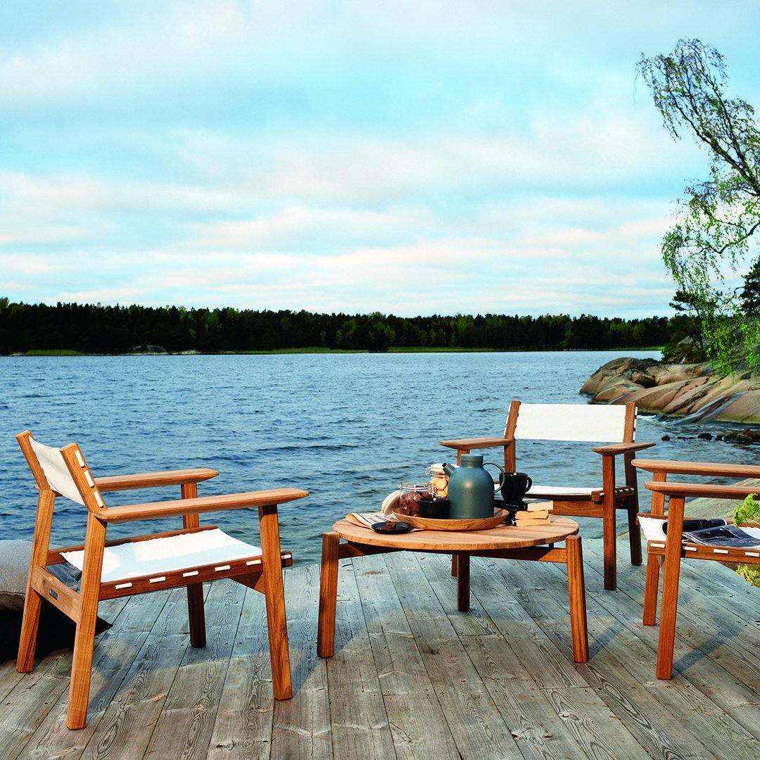"Light wood outdoor furniture with white cushions, set against a serene lakeside backdrop and soft blue sky."