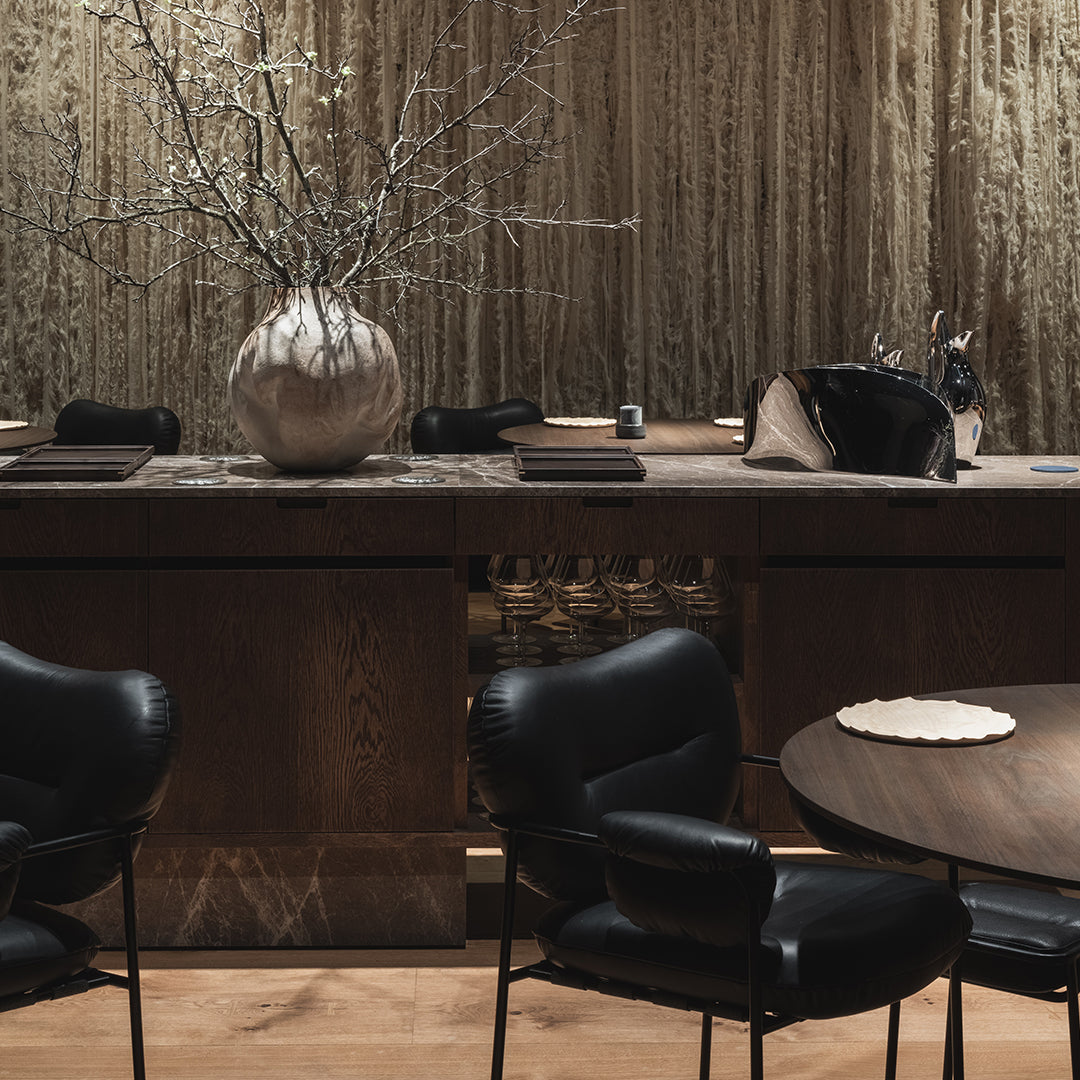 Black chairs with curved backs, earthy wood tones, and neutral textures create a modern, minimalist dining space aesthetic.