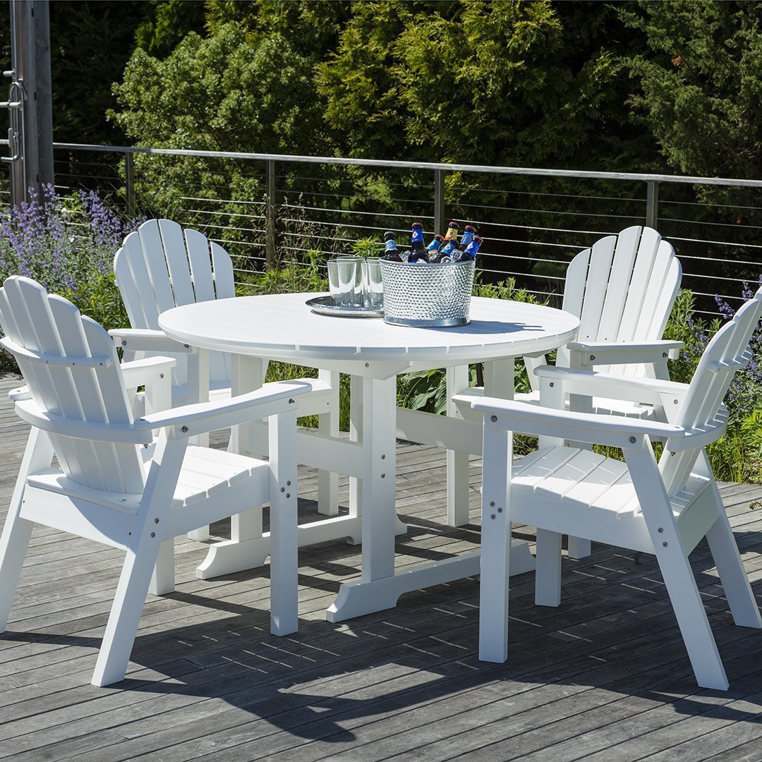 White outdoor dining set with circular table and Adirondack chairs against a natural green backdrop.