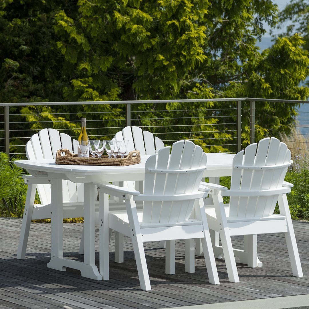 White Adirondack chairs and table set on a wooden deck, surrounded by lush green trees and a clear blue sky.