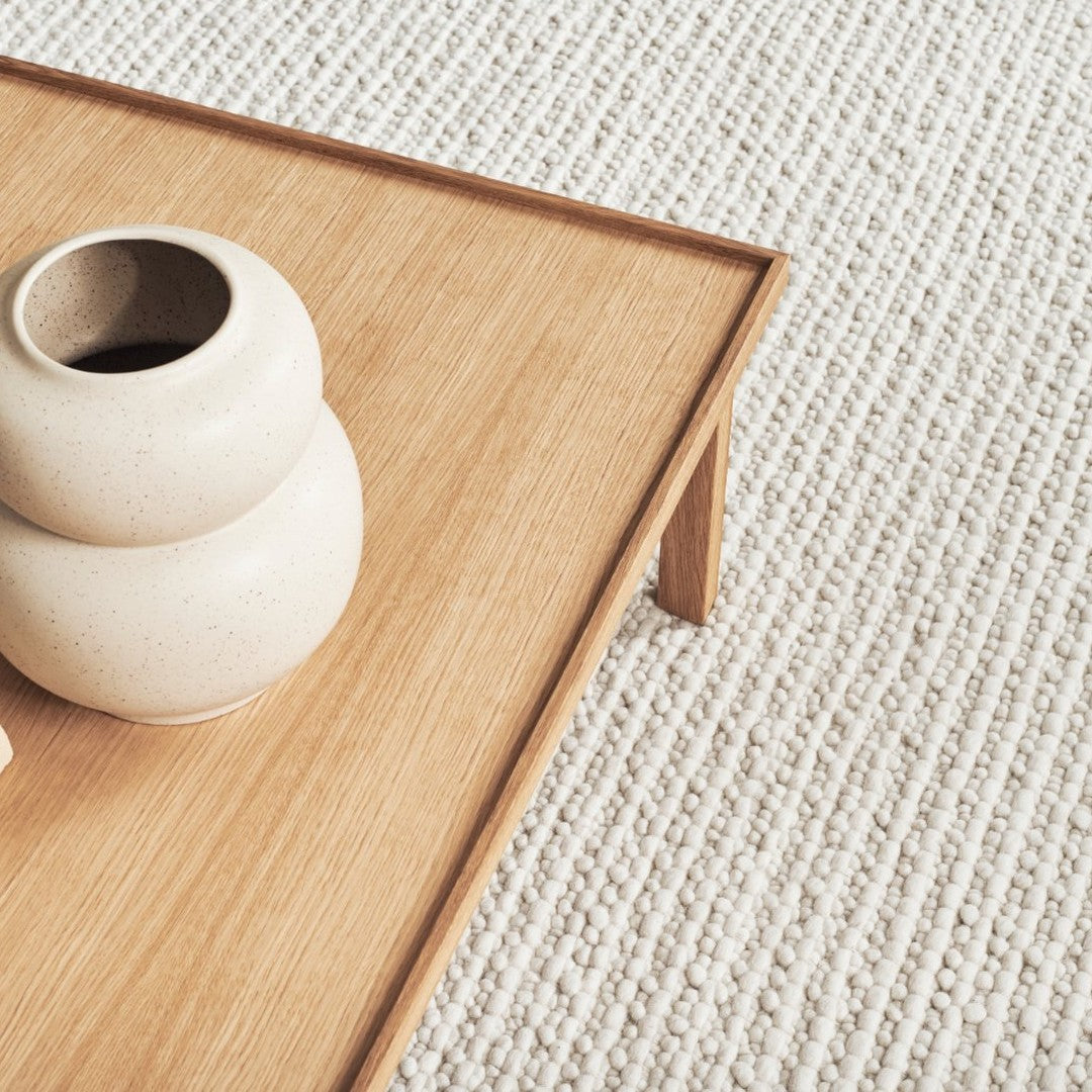 Light beige textured rug with natural wood table and cream ceramic vases. Subtle neutral tones for a cozy aesthetic.