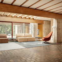 Modern living room with a beige couch, rust-colored accent chair, light gray rug, and wooden beams overhead.