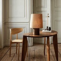 Wooden table with a natural finish, paired with a beige lampshade and a light oak chair, set against neutral walls.