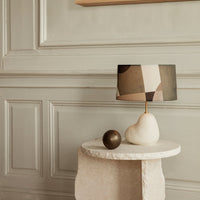Geometric-patterned lampshade in neutral tones atop a smooth white base, with a dark spherical accent on a light stone table.