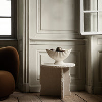 White textured bowl on a light-toned pedestal table, against a muted gray wall, with warm brown furniture accents.