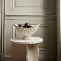 Cream ribbed bowl with decorative dark and light orbs, set on a textured stone pedestal. Modern, elegant design.