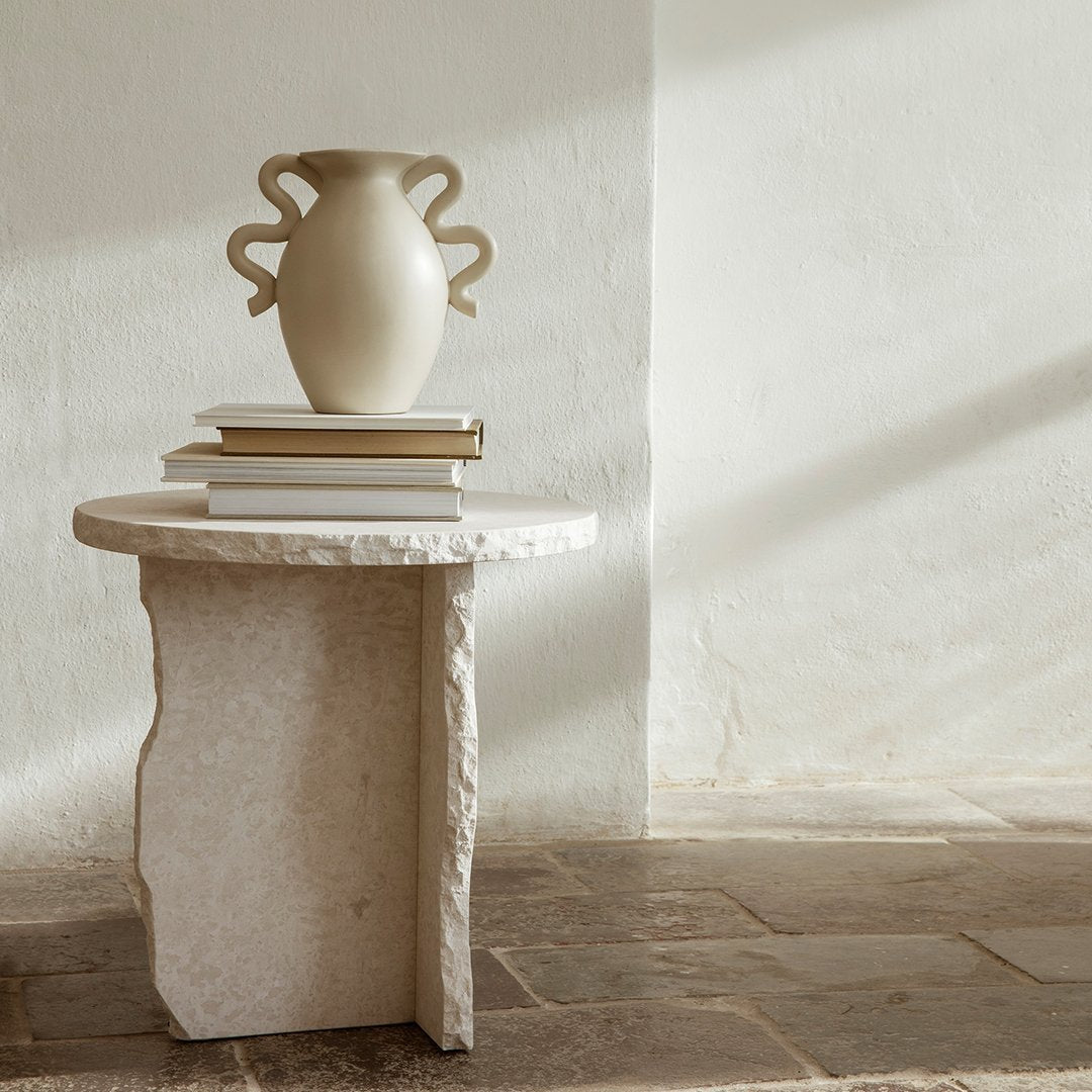 Neutral beige vase atop a textured stone table, against a soft, light-colored wall with gentle shadows.