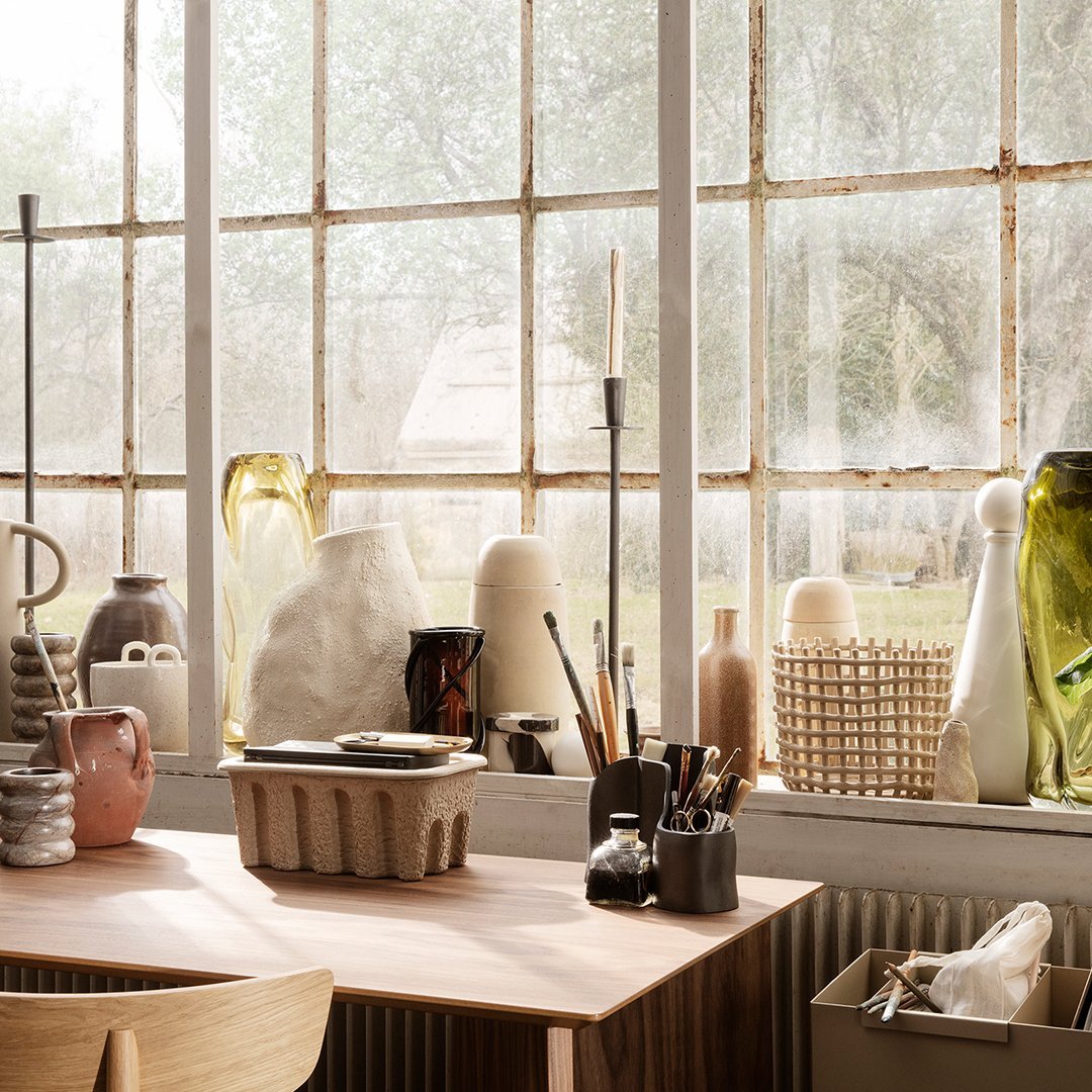 "Array of textured vases in soft pastels, greens, and clear glass, showcased on a wooden table by a sunlit window."