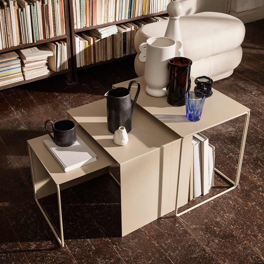 "Set of three nesting tables in beige with sleek lines, complemented by black and blue glassware on a warm wooden floor."
