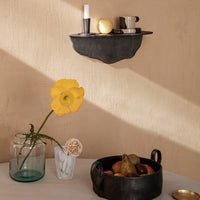 Black wall shelf with natural textures, vibrant yellow flower, earthy tones, and assorted fruits in dark bowl.