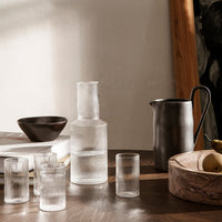 Clear textured glass pitcher and tumblers with a dark ceramic bowl, set on a wooden table. Natural earthy tones.