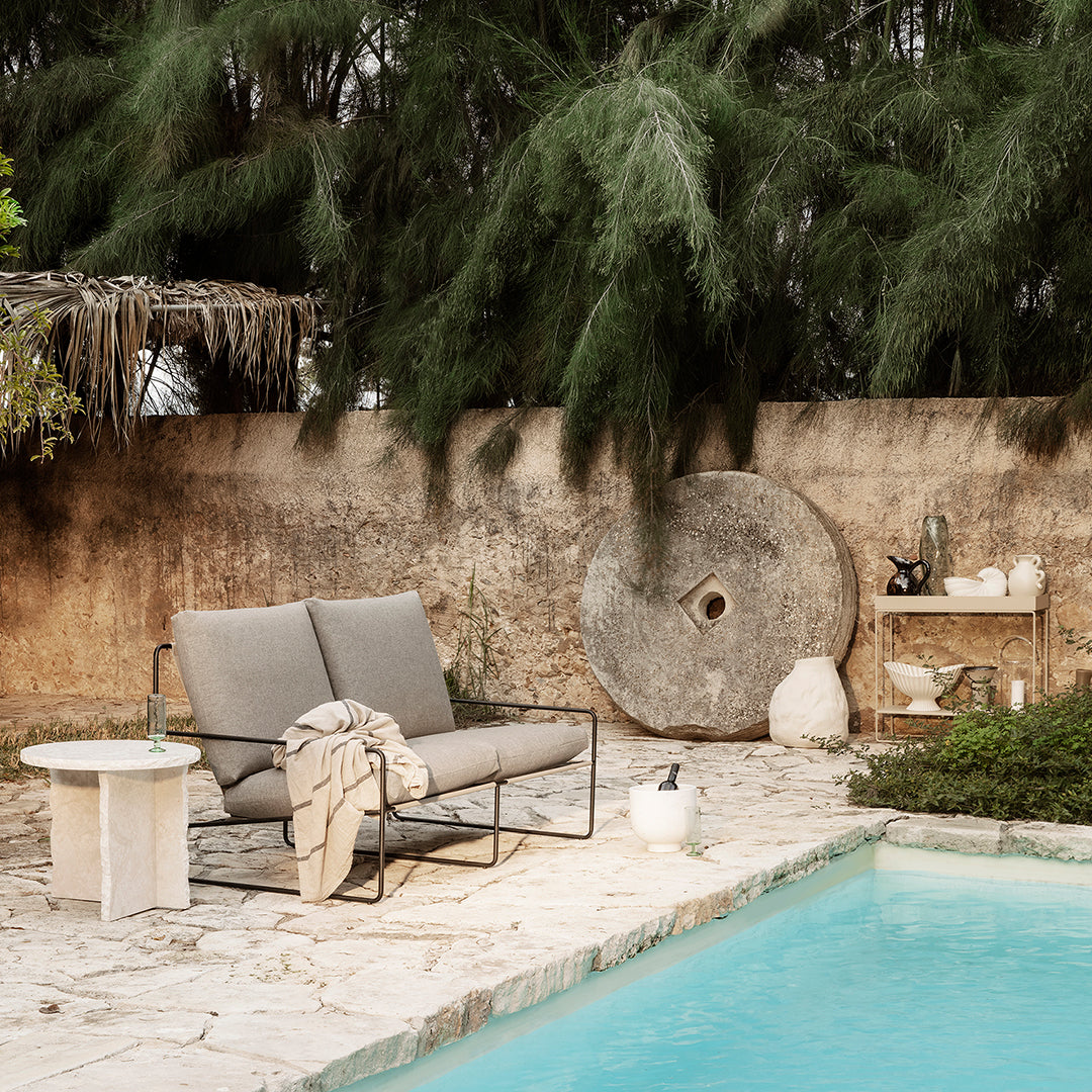 Gray outdoor sofa with a neutral throw, surrounded by earthy tones and lush greenery by a turquoise pool.
