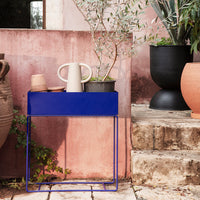 Vibrant blue metal console table against a textured pink wall, surrounded by earthy tones of pots and greenery.