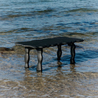Black scalloped table with curved legs on a sandy beach, contrasting against clear blue water.