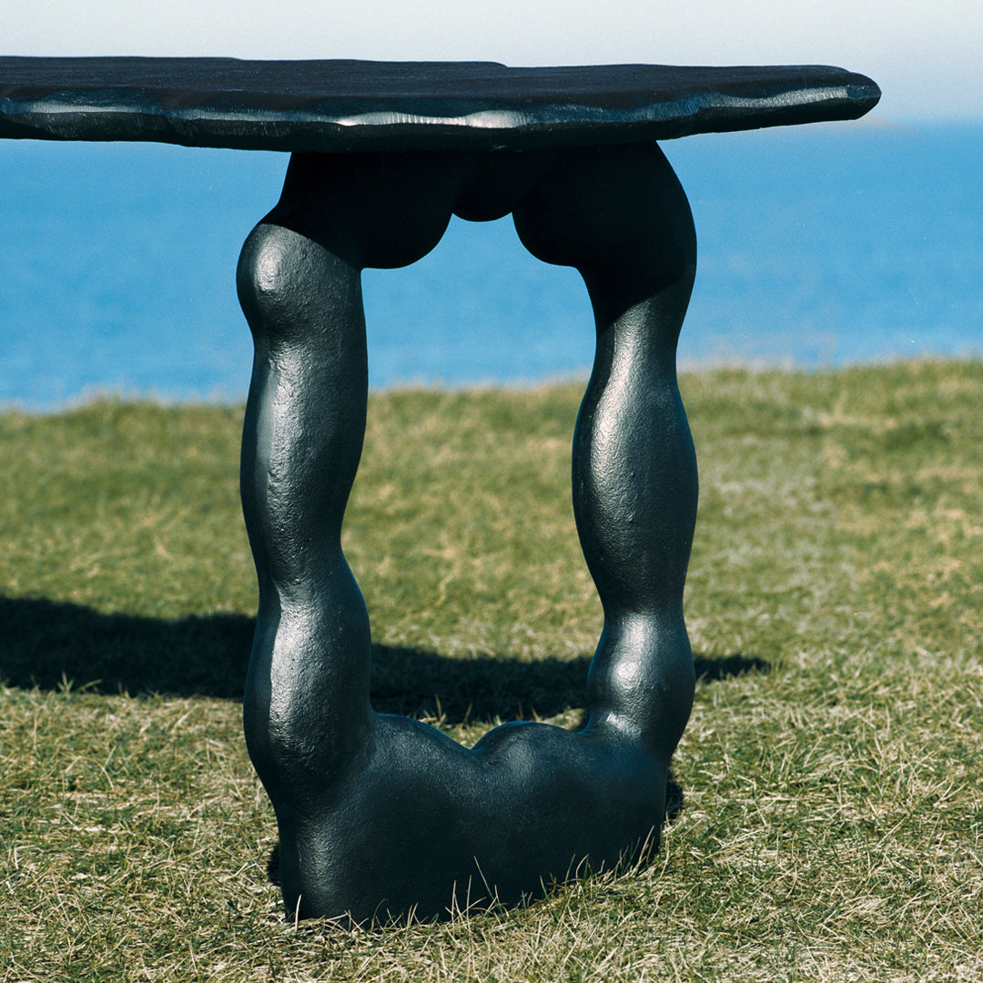 Black sculptural table base with a smooth, organic shape, set against a grassy surface and ocean backdrop.
