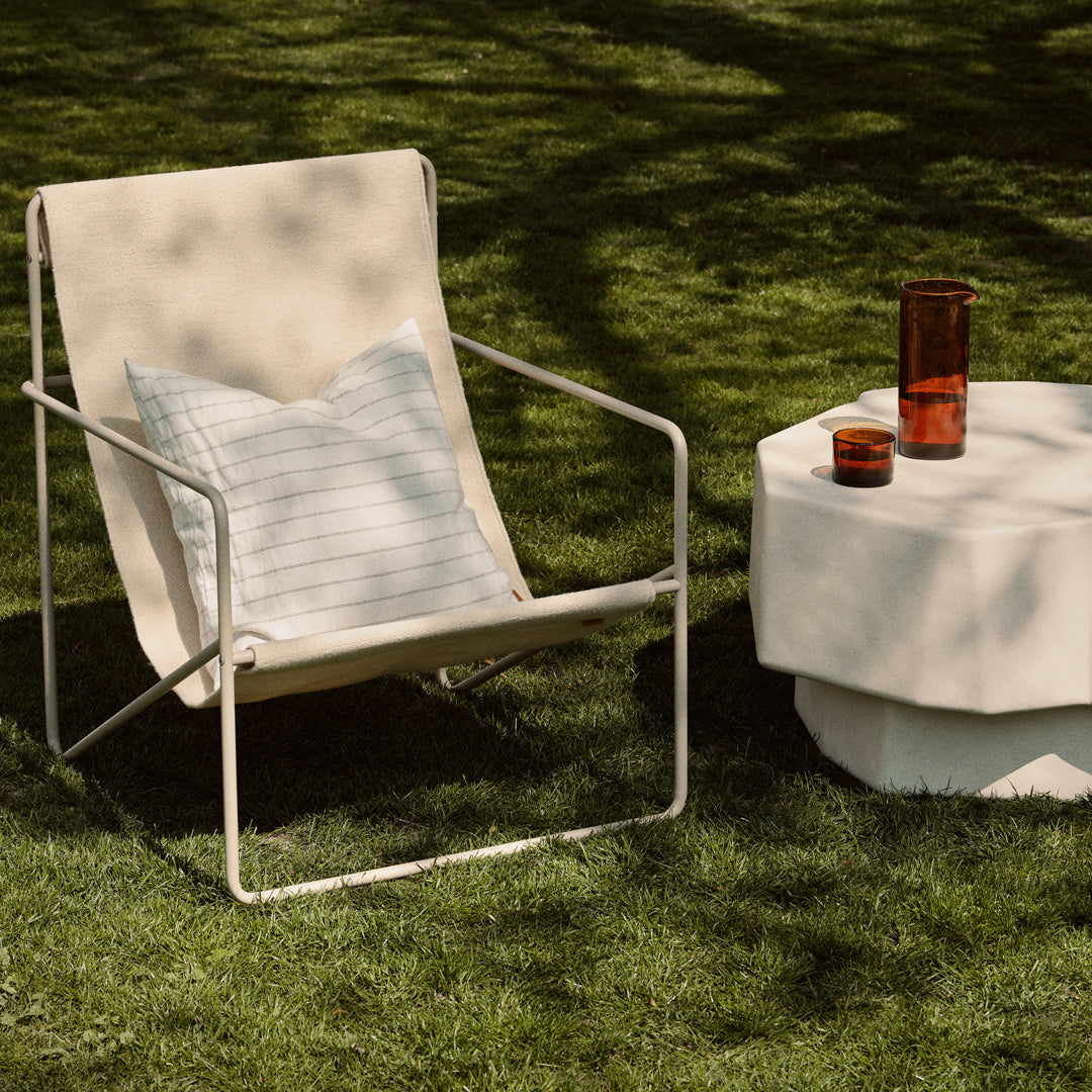 Cream canvas chair with a striped cushion, minimalist white table, and amber glass accents on green grass.