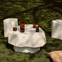 Modern white outdoor table with organic shape, complemented by amber glass decor on lush green grass. Perfect for stylish gatherings.