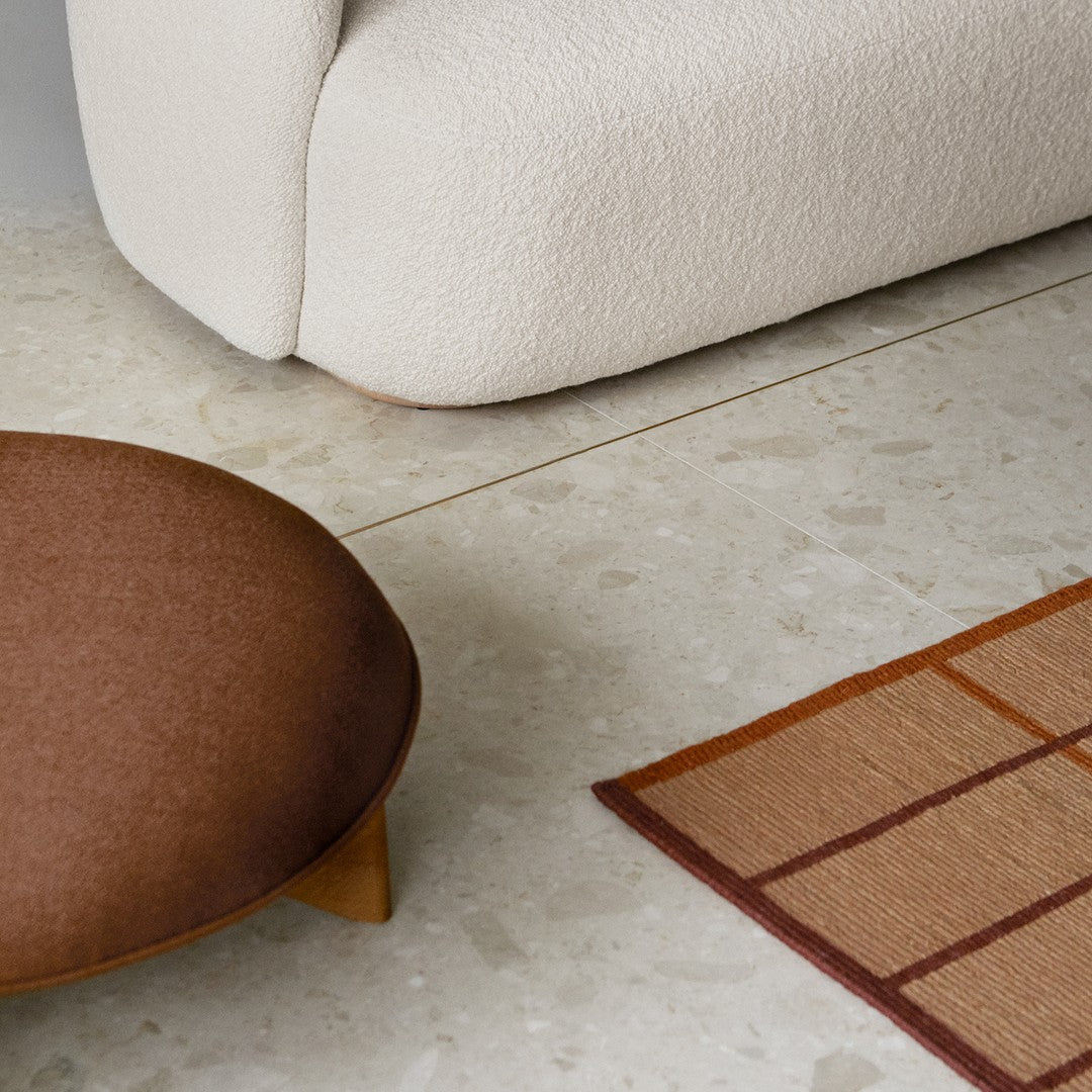 Brown round ottoman and geometric patterned rug in warm tones against a light, textured floor.
