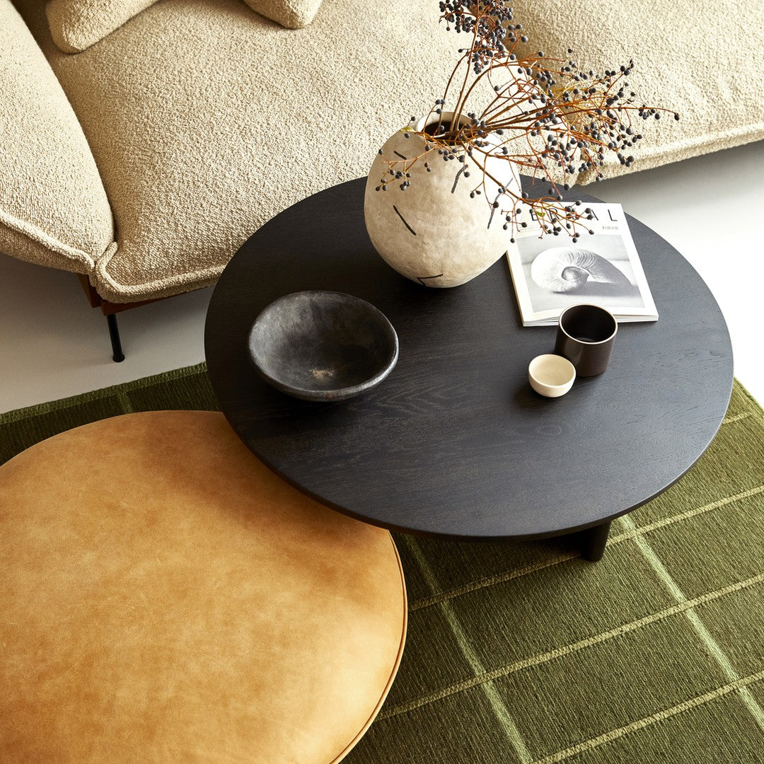 Black round coffee table on green plaid rug, with tan circular stool and white textured vase. Minimalist decor.
