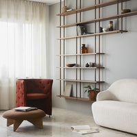 "Red tufted armchair, beige curved sofa, and wooden shelving against a light gray wall in a modern living room."