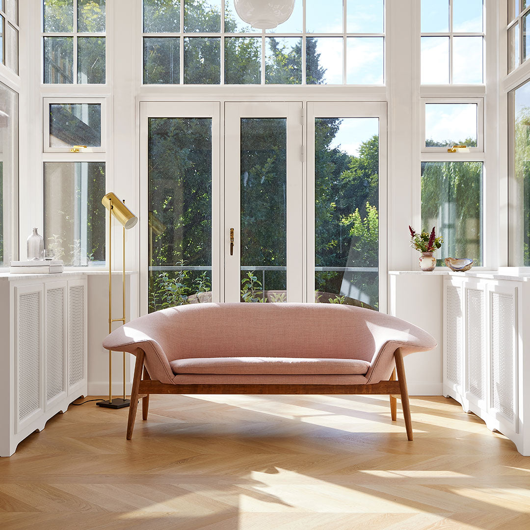Soft pink fabric sofa with a sleek wooden frame, set against bright, natural light and modern, airy decor.