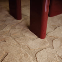 Beige area rug with textured stone pattern, featuring soft, warm tones and contrasting glossy red table legs.