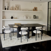 Modern black dining table with white chairs on a light rug, set against a minimalist white background.